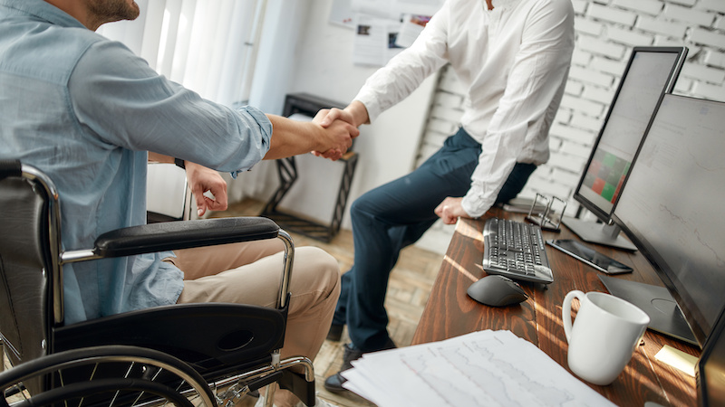 Cropped view of male office worker in a wheelchair shaking hand to his colleague while working together in the creative office. Teamwork. Disability and handicap concept. Business meeting