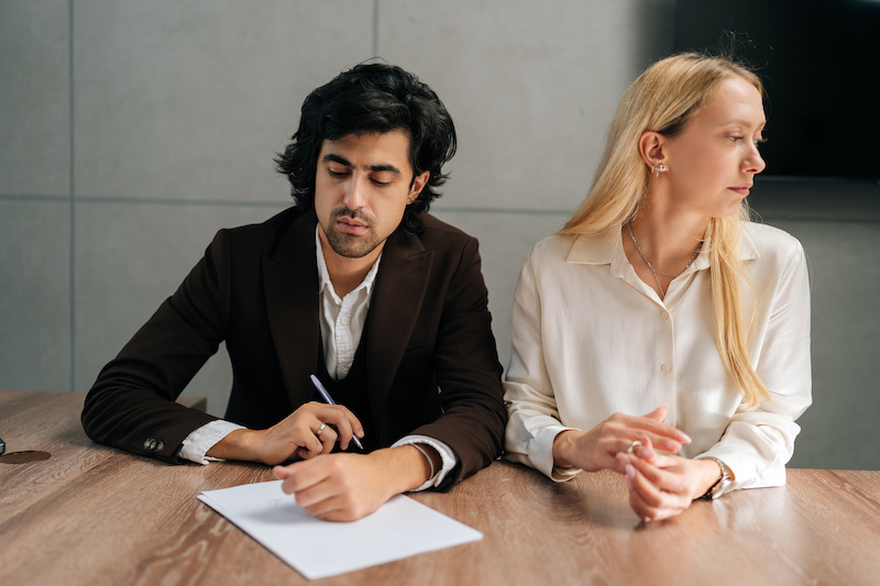 Portrait of sad spouses couple signing decree papers getting divorced in lawyers office at desk. Unhappy married man and woman filing divorce, shares or mortgage assets with attorney.