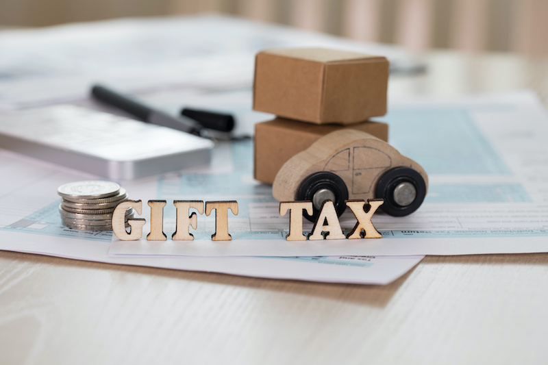 Word GIFT TAX composed of wooden letters. Coins, papers, pen, wooden car, carton boxes in the background. Closeup