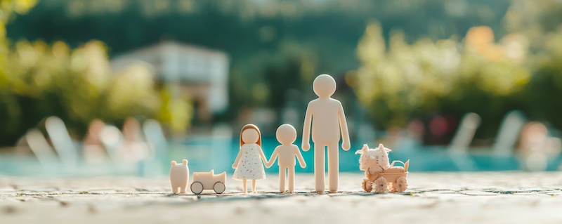 Wooden figurines of a family and pets capturing a heartwarming moment outdoors, blurred background with greenery and a pool.