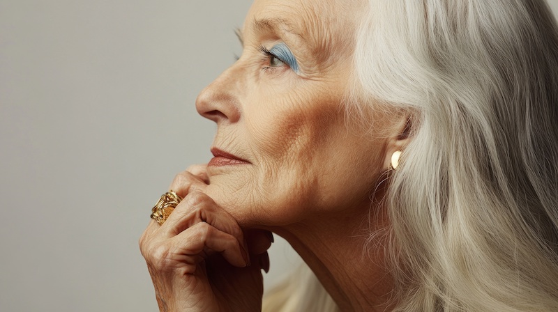 A woman with blue eyes and gray hair is wearing a gold ring and earrings. She is looking down at her hands