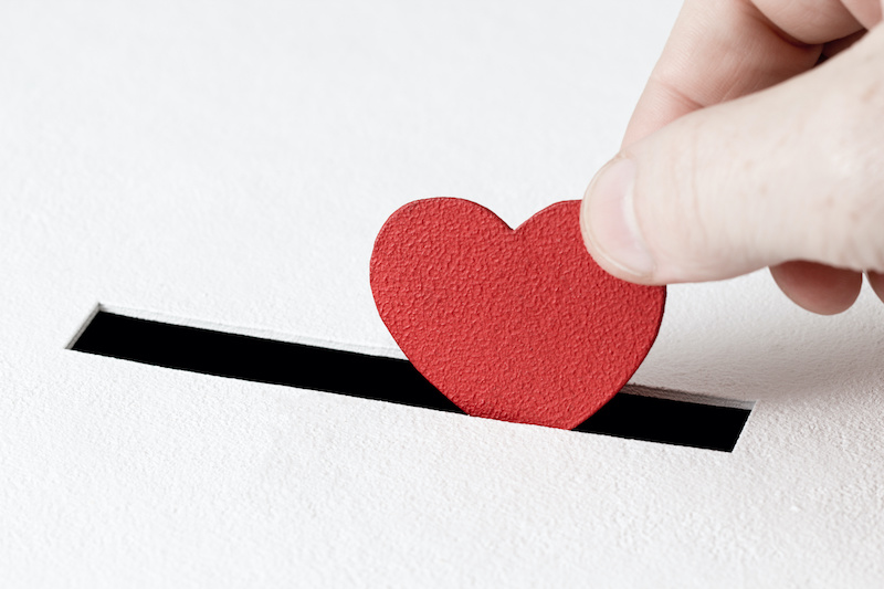 Hand of man inserts heart symbol into hole in donation box on white background with copy space. Concept of donor, life-saving or kindness