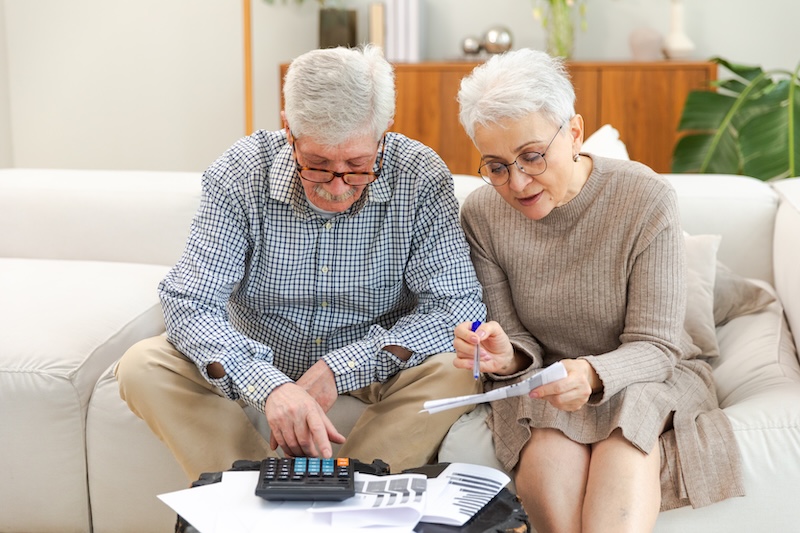 Middle aged senior couple sit with laptop and paper document. Older mature man woman reading paper bill pay online at home managing bank finances calculating taxes planning loan debt pension payment