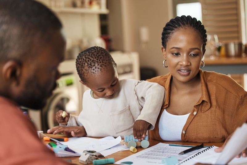 Busy, tired and multitask mother working while taking care of her child at home. African american entrepreneur or freelancer analyzing paperwork with her husband while holding her busy and cute son.