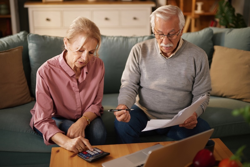An older man and woman sit on a sofa at home, examining paperwork and using a calculator and laptop. They appear to be managing their finances or discussing insurance options.