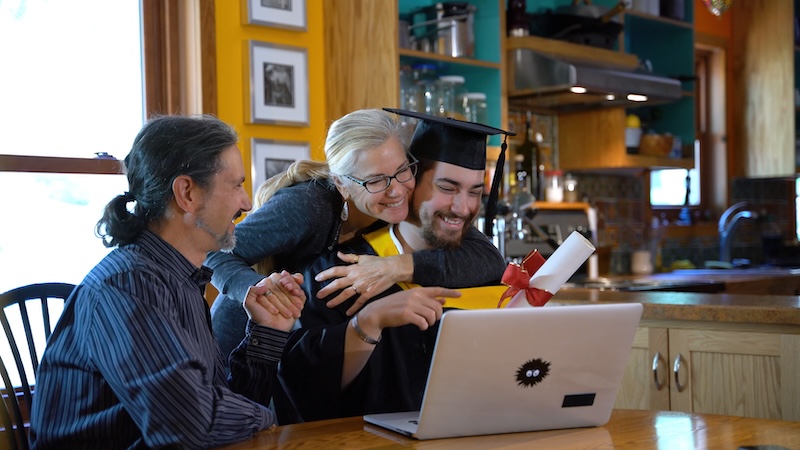 Young man moves tassel to other site for virtual commencement ceremony for graduation, then smiles and everyone hugs.