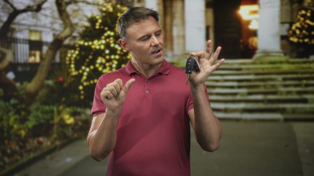 Man holds car keys in right hand and makes a pinch gesture with left while standing before building steps and festive lights; homecoming confidence.