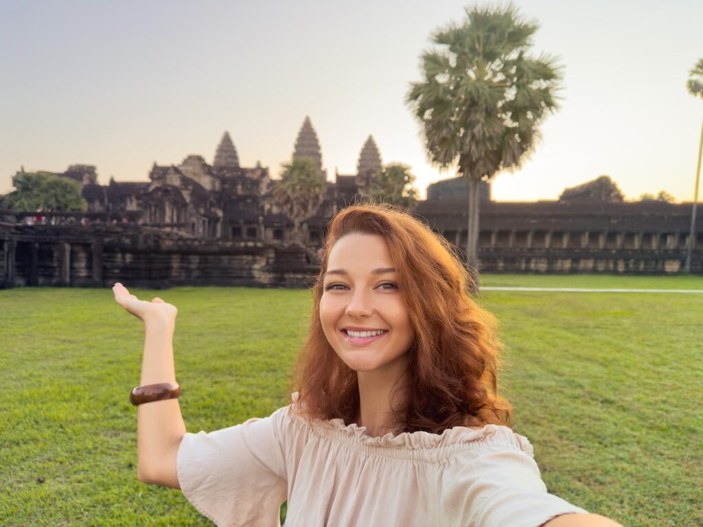 Cheerful young female traveler poses for a selfie and gestures toward Angkor Wat at sunrise. A high quality stock image ideal for travel blogs, tourism campaigns, and cultural exploration themes.